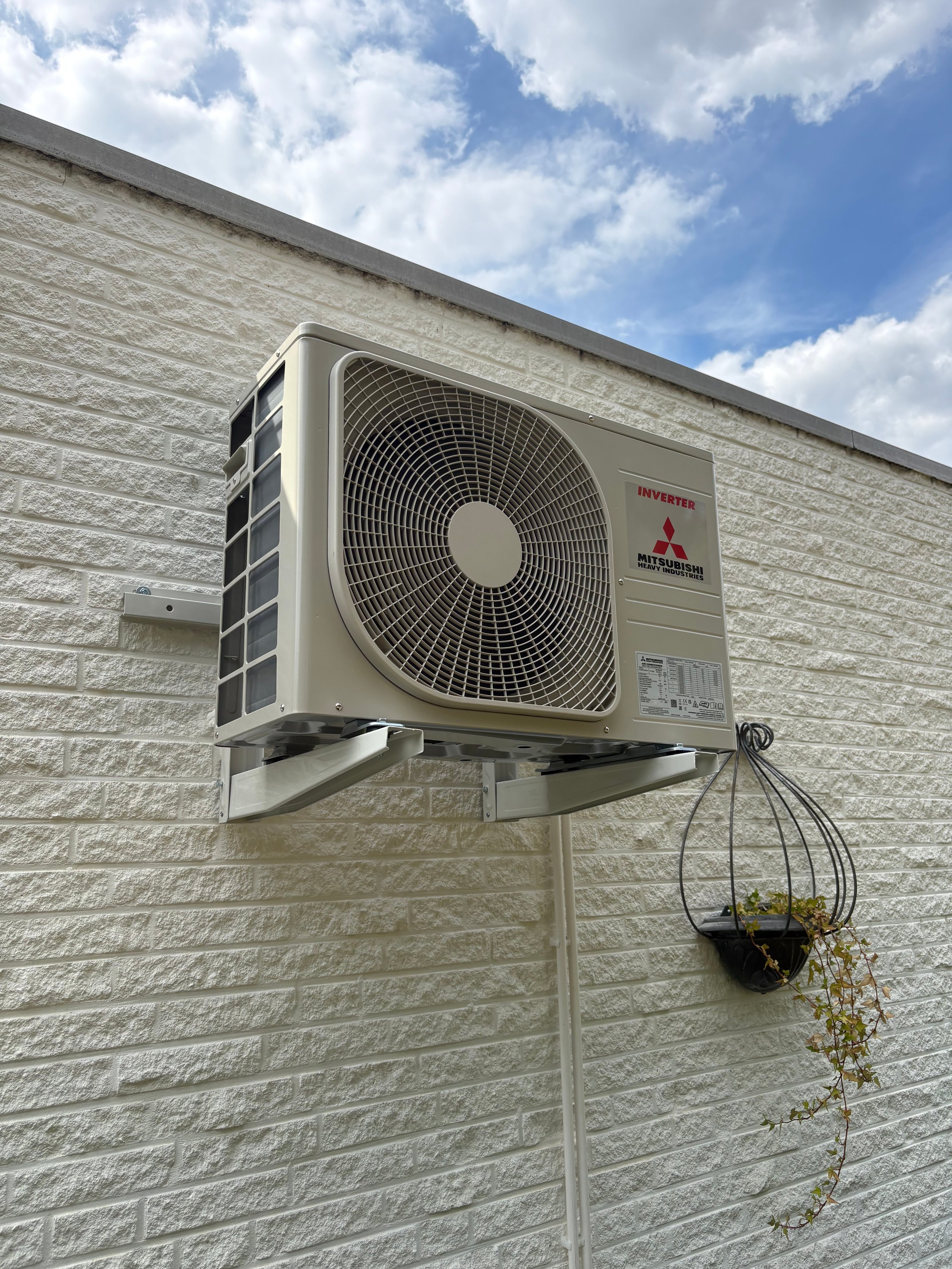 Mitsubishi air conditioning unit mounted on white brick wall with cable and hanging plant below it