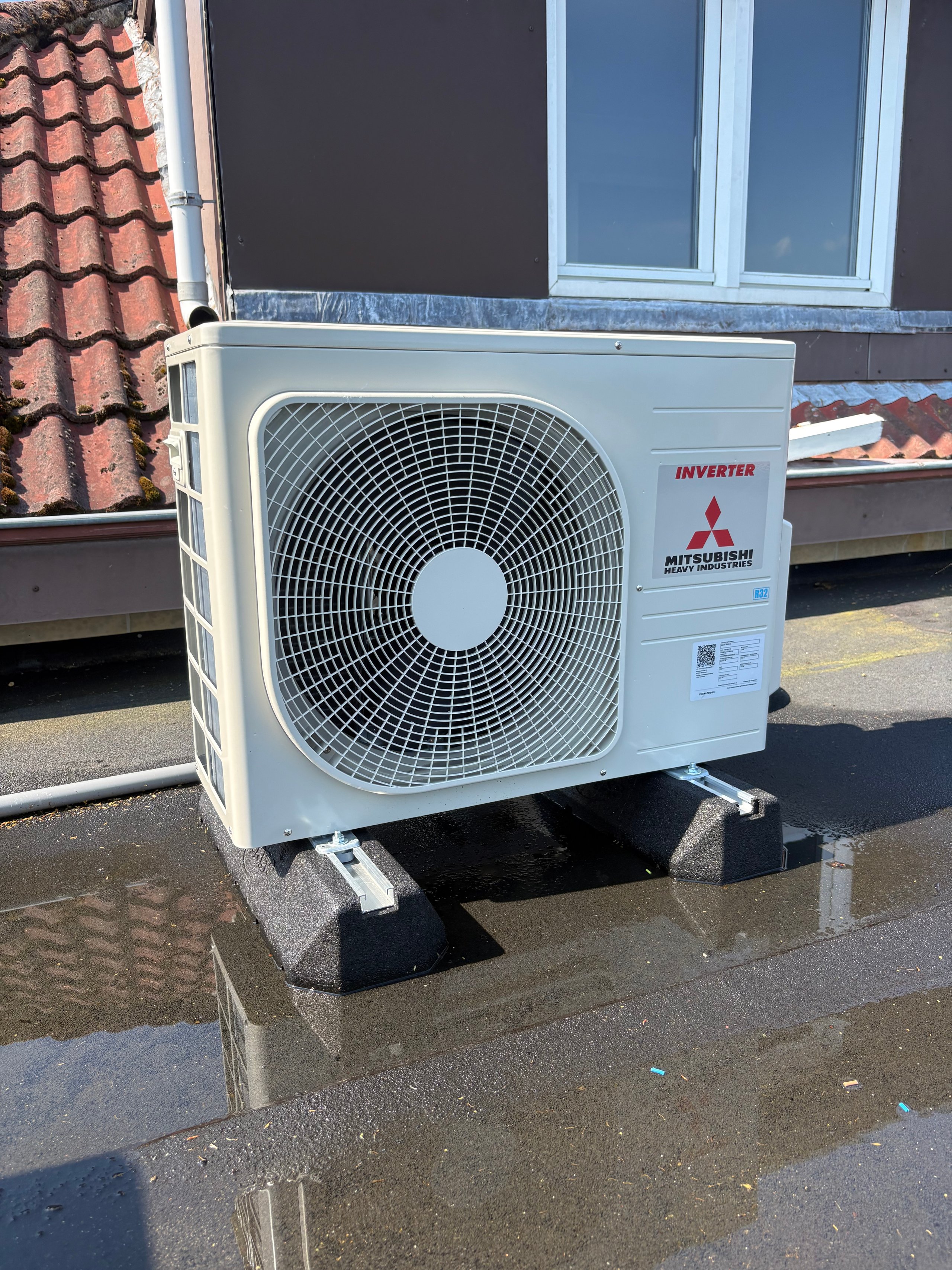 White Mitsubishi inverter heat pump outdoor unit mounted on a wet roof with terracotta tiles visible