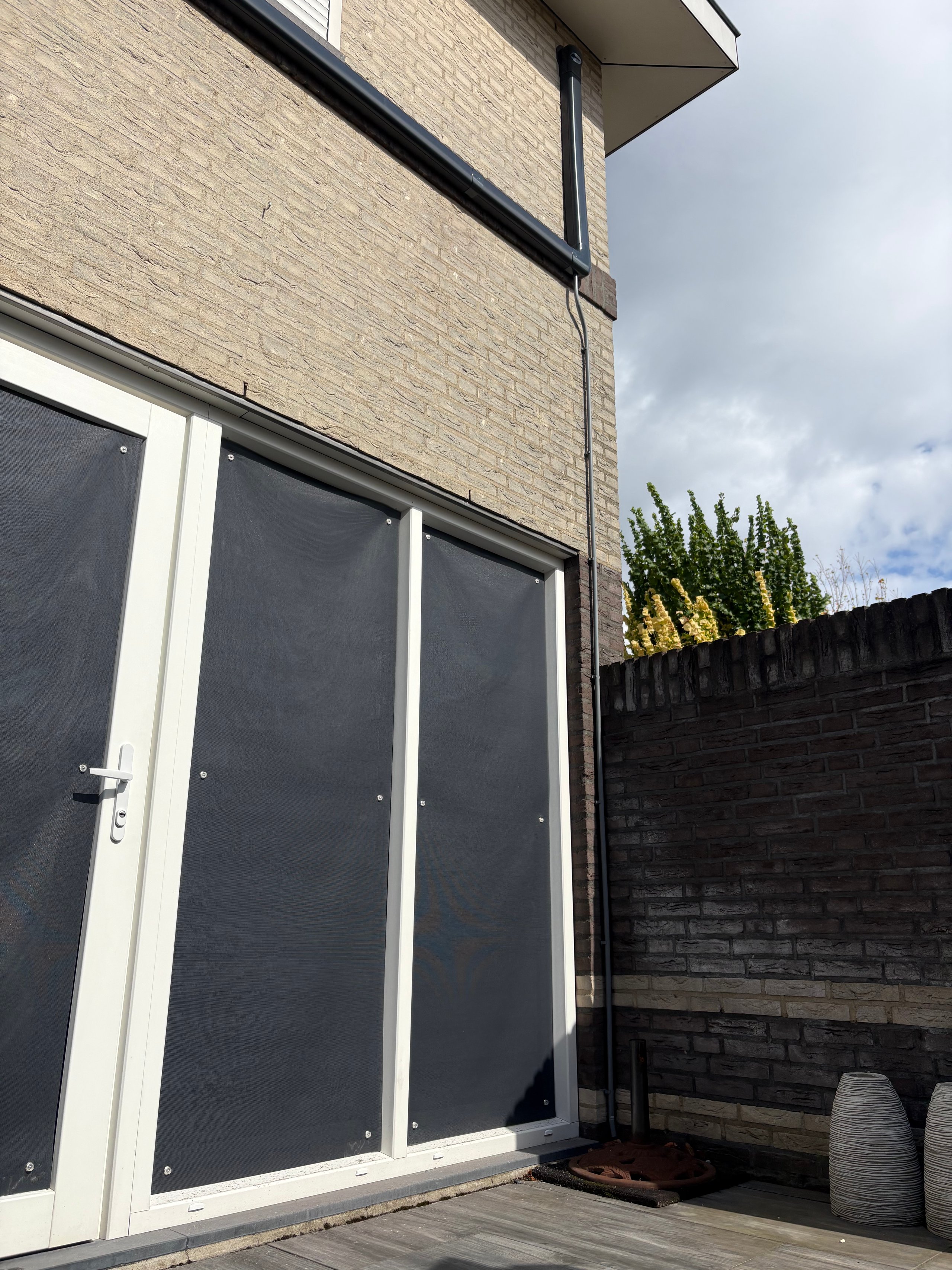 Modern residential exterior with tan brick wall, white-framed glass doors, dark wooden fence, and patio area under cloudy sky