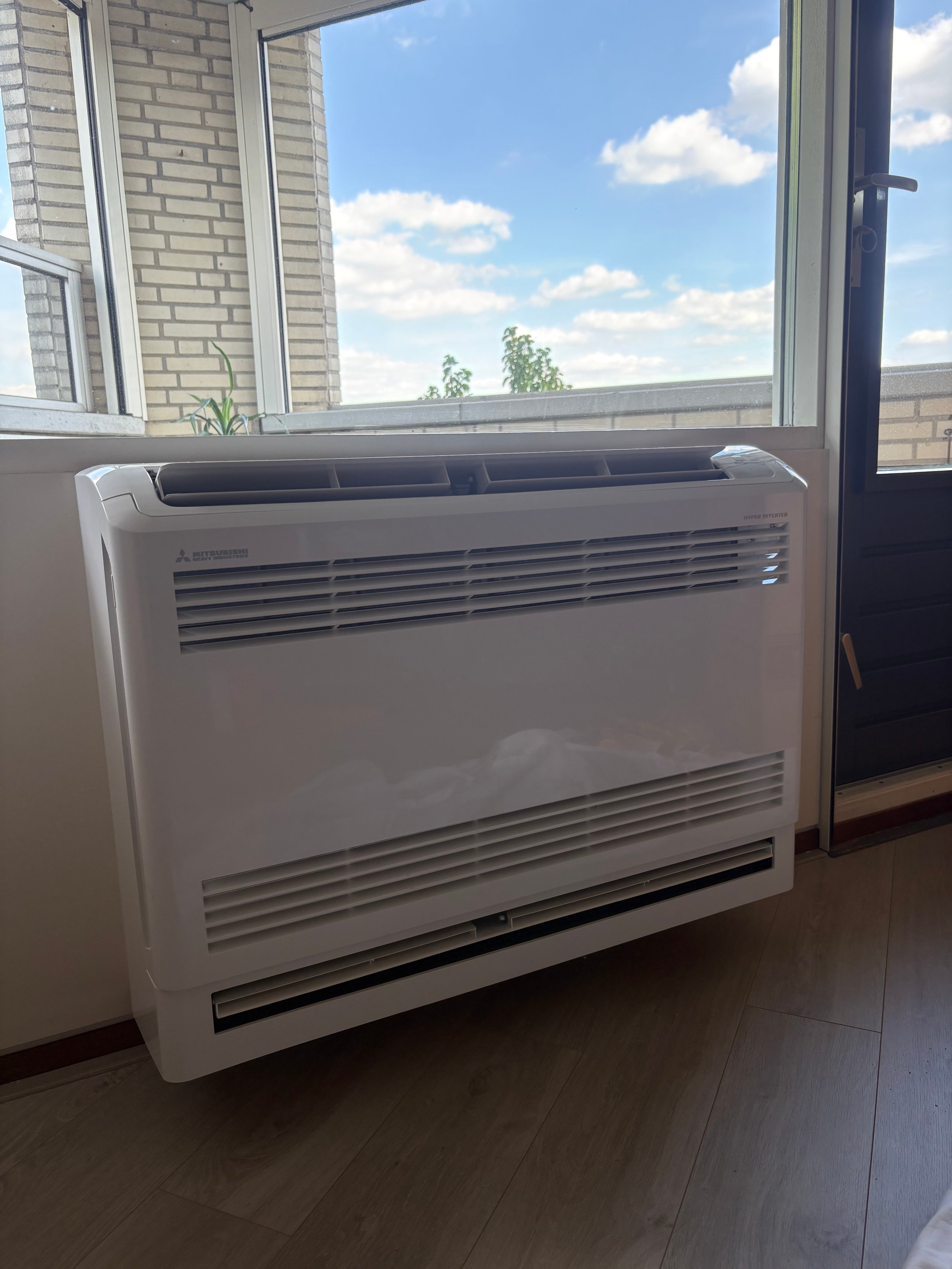 White floor-mounted air conditioning unit positioned near a window overlooking a residential area