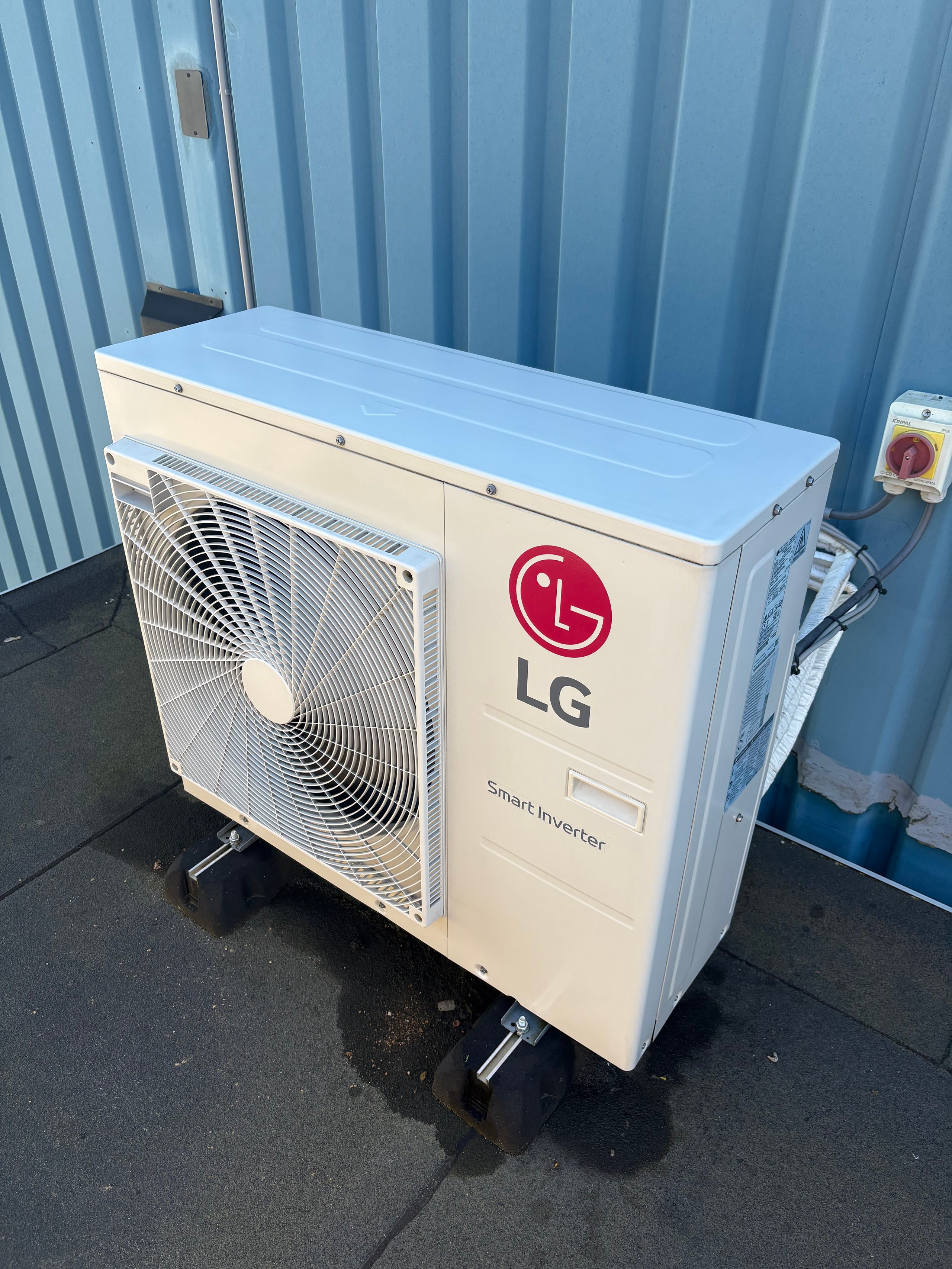 LG air conditioning unit outdoor compressor with white casing and fan mounted on wheels against corrugated metal wall