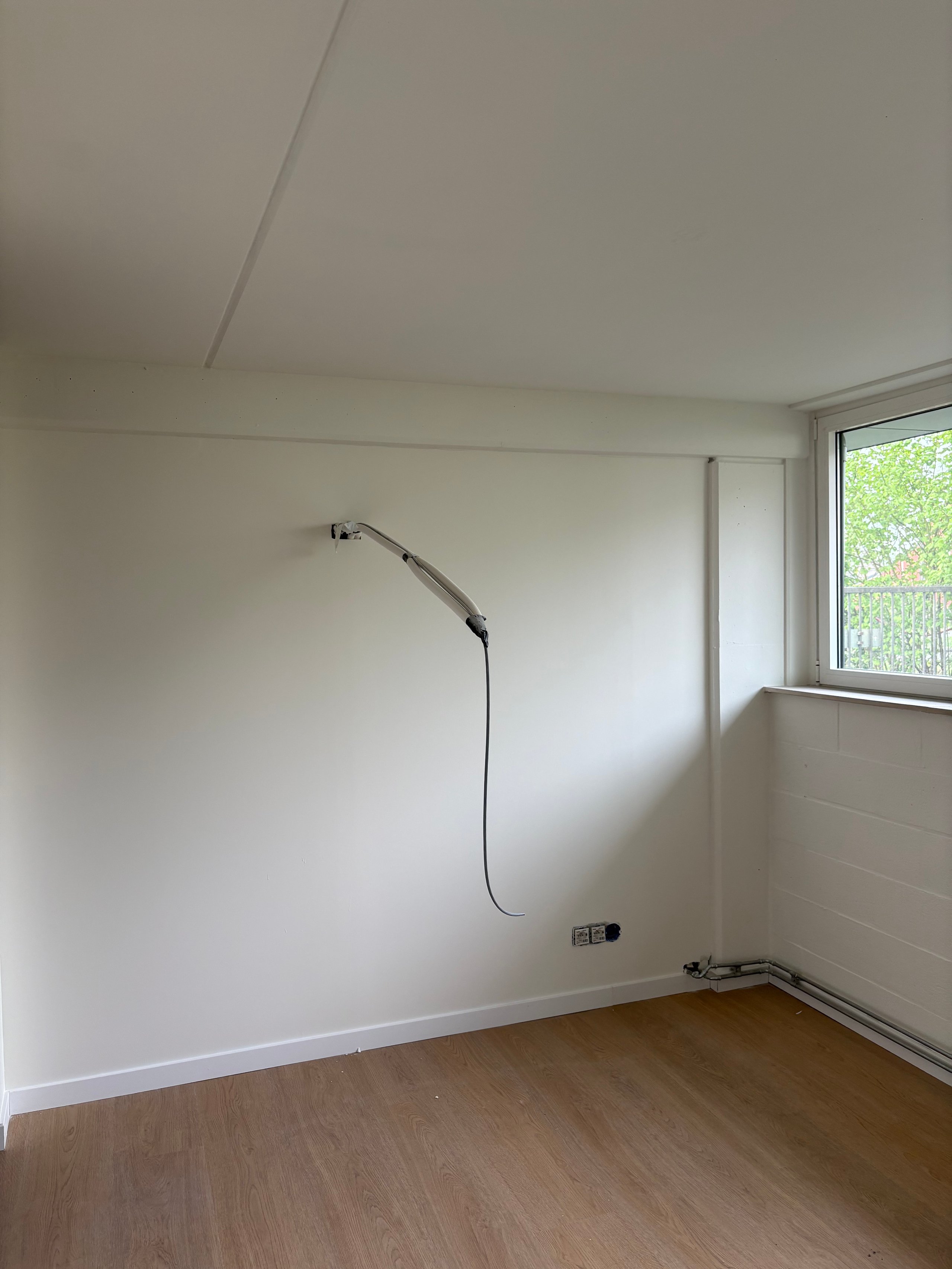 Empty room with white walls, wooden floor, mounted wall lamp, window with green view, and power outlets visible
