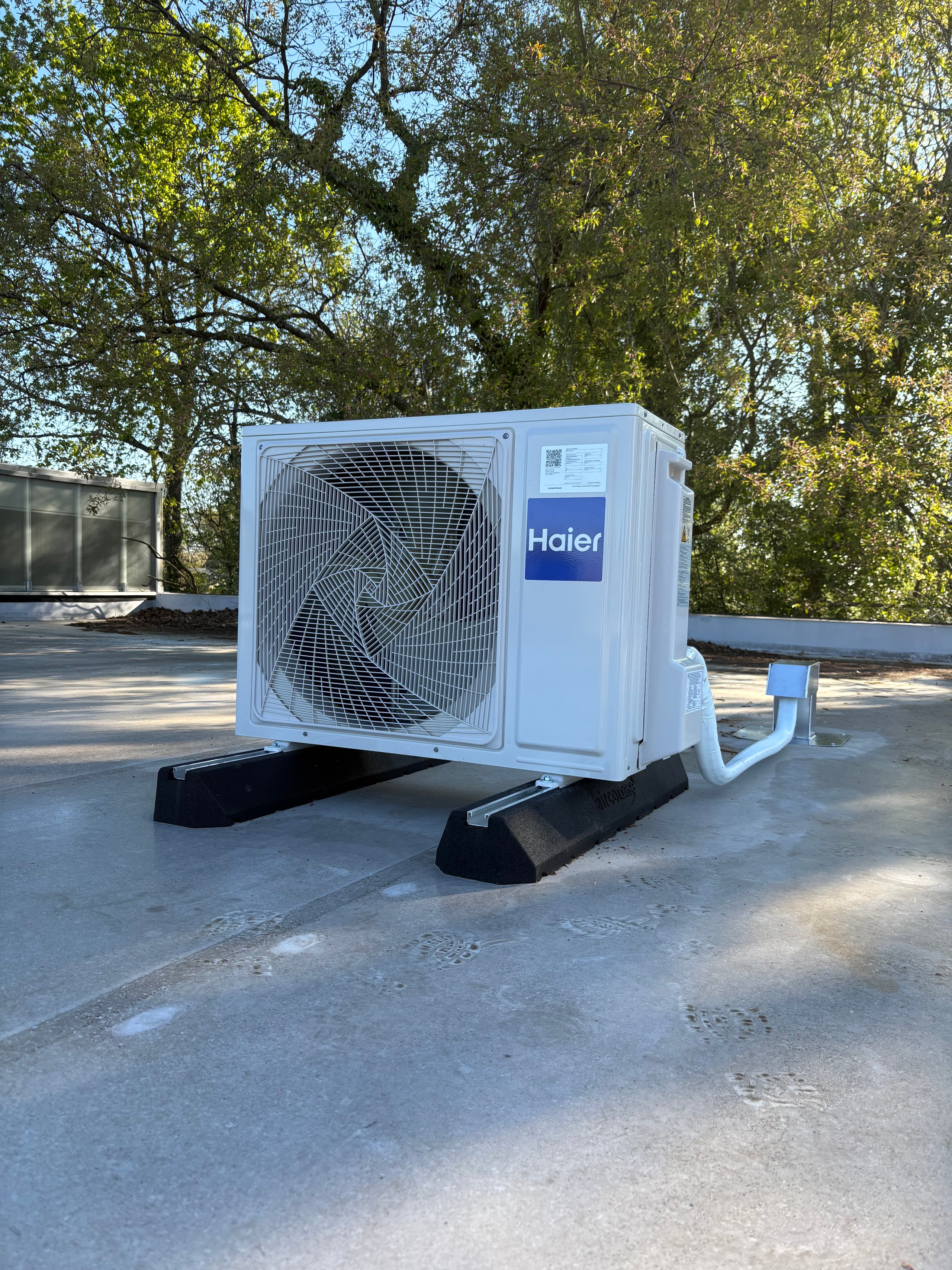 Haier air conditioning unit outdoor compressor installed on concrete pad under trees on rooftop