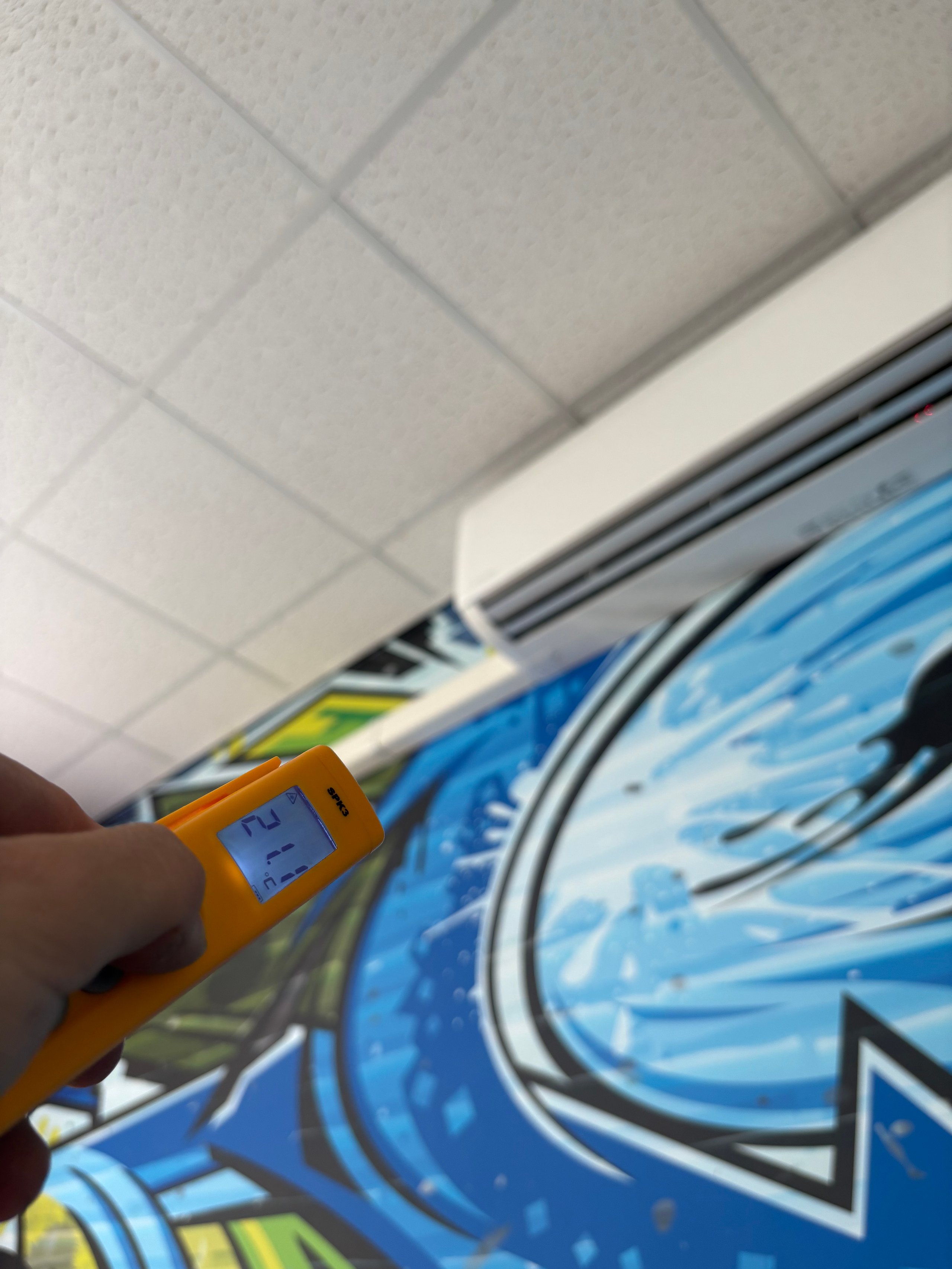 Hand holding yellow digital thermometer pointing at ceiling tiles and air conditioning duct in indoor setting