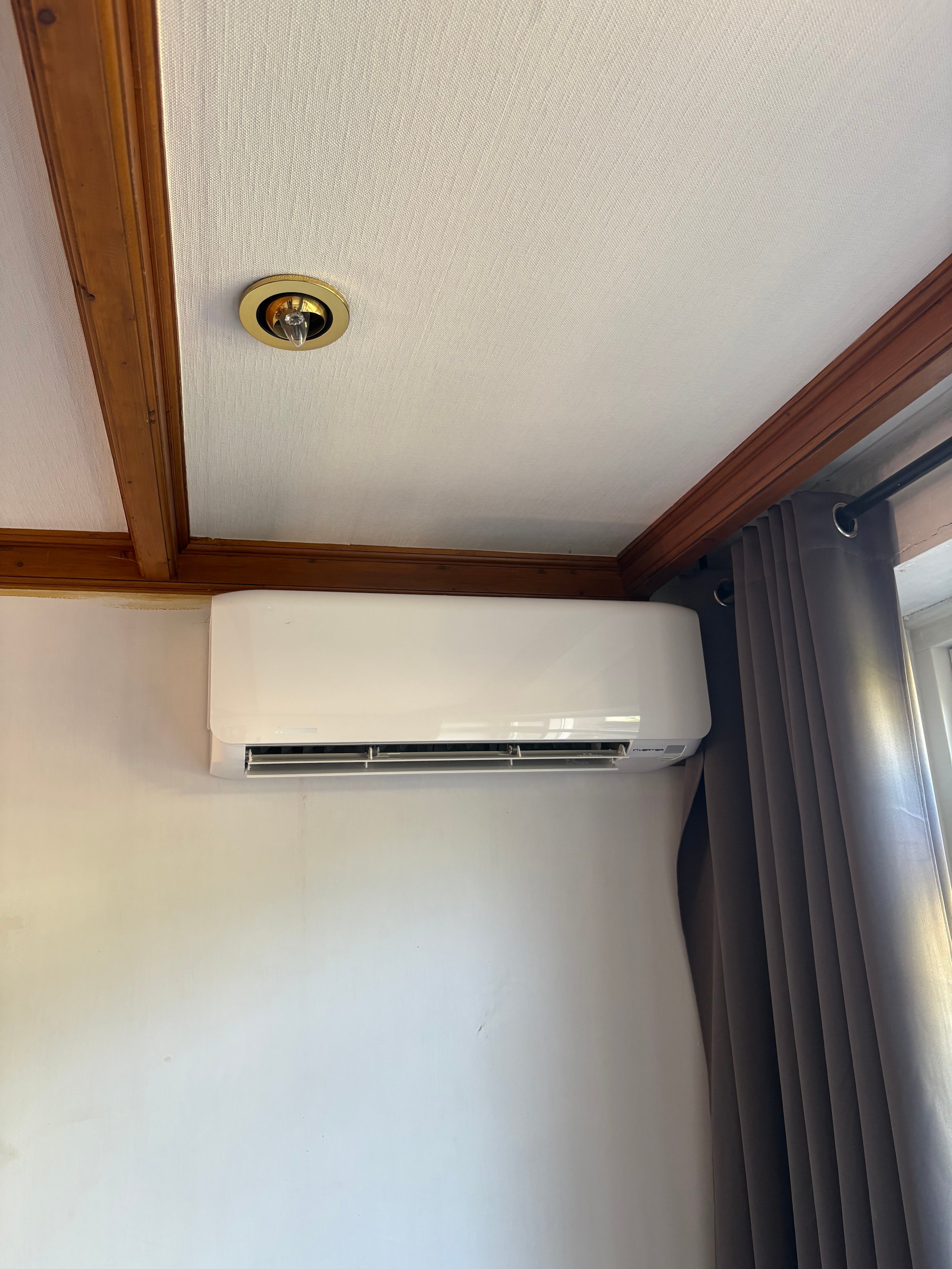 White wall-mounted air conditioning unit in room with wooden ceiling trim and dark curtains