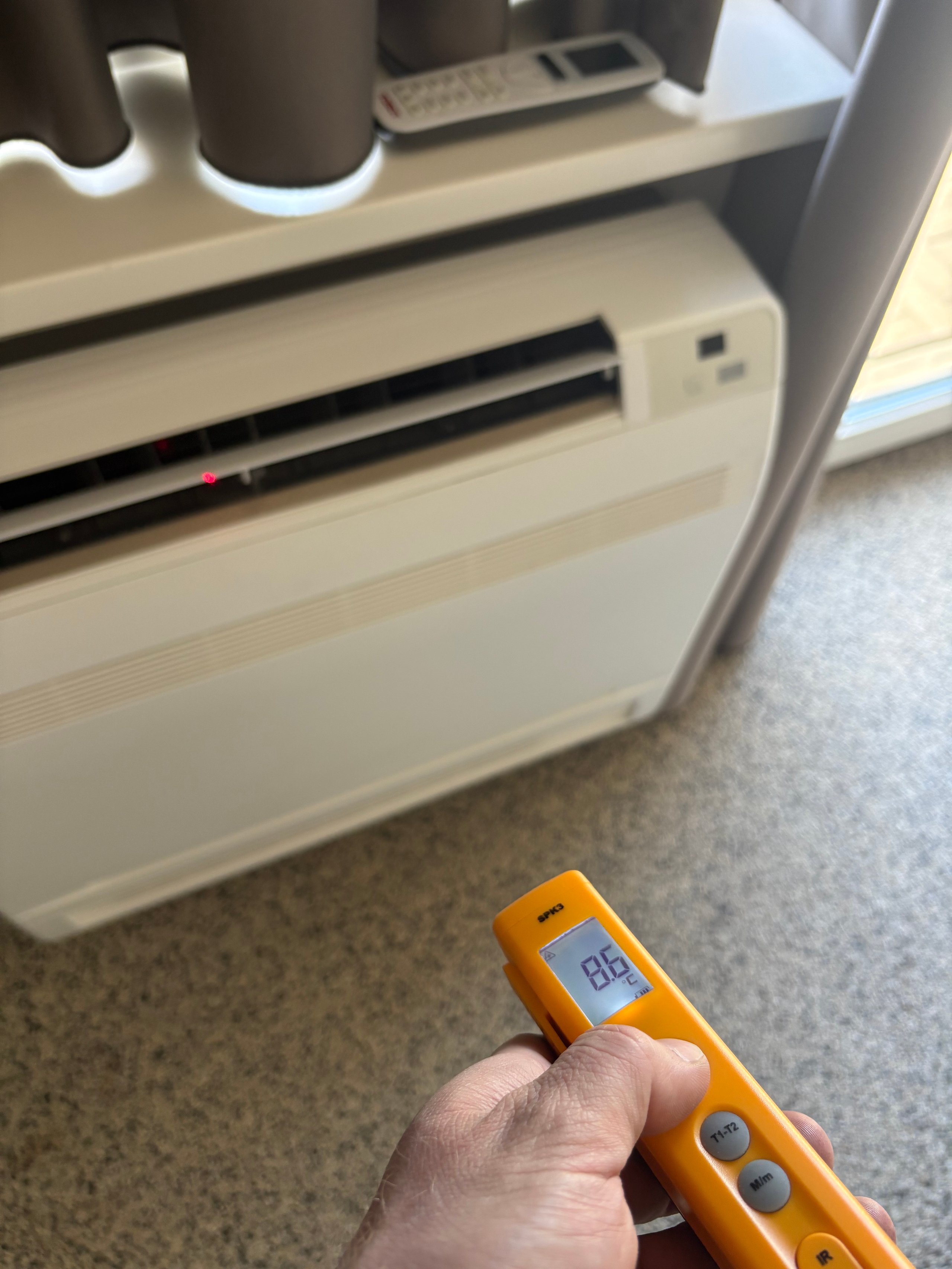 Hand holding orange digital thermometer displaying temperature reading in front of white portable air conditioning unit