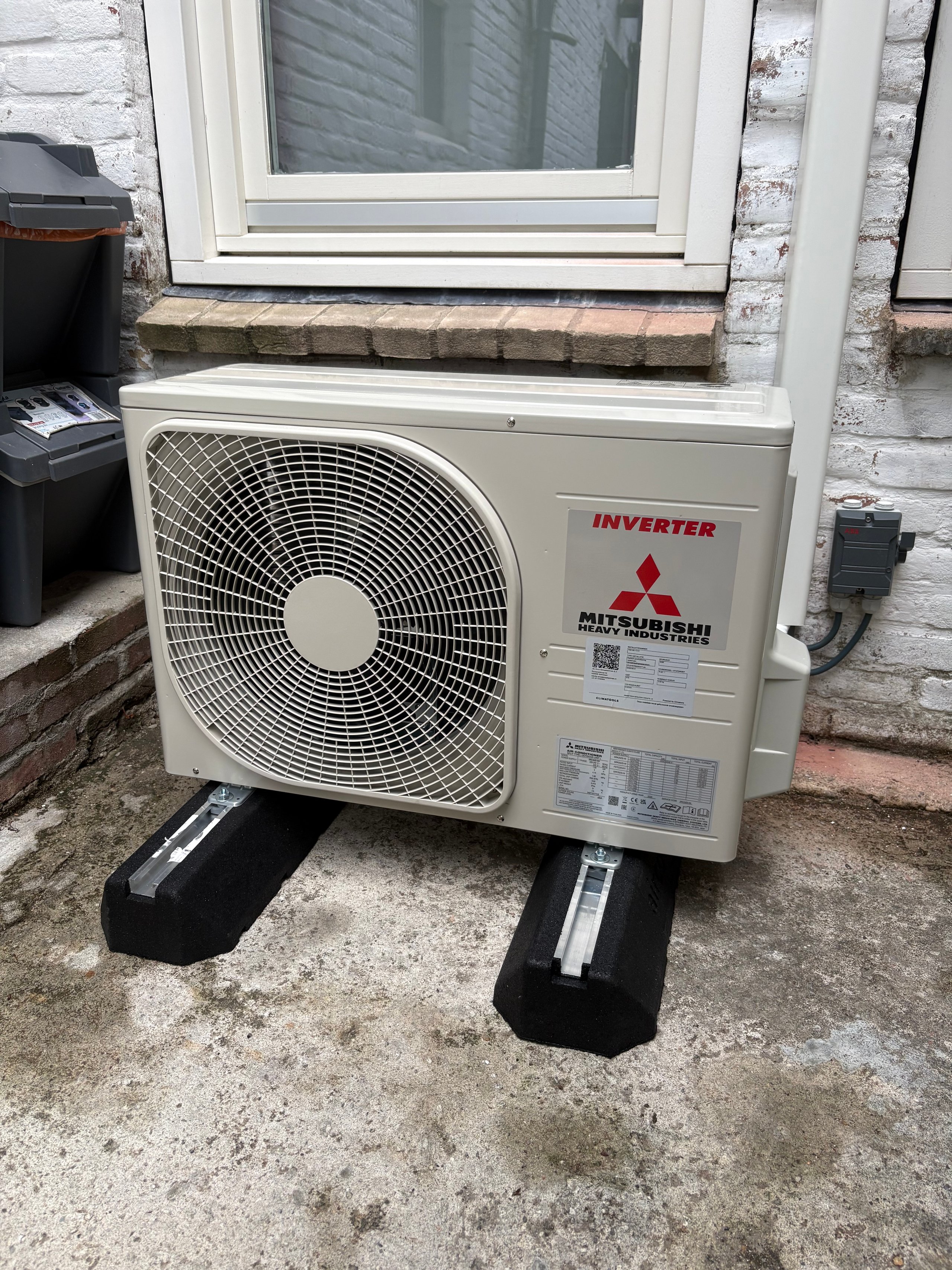 Mitsubishi inverter air conditioning outdoor unit mounted on black stands against a brick wall below a window