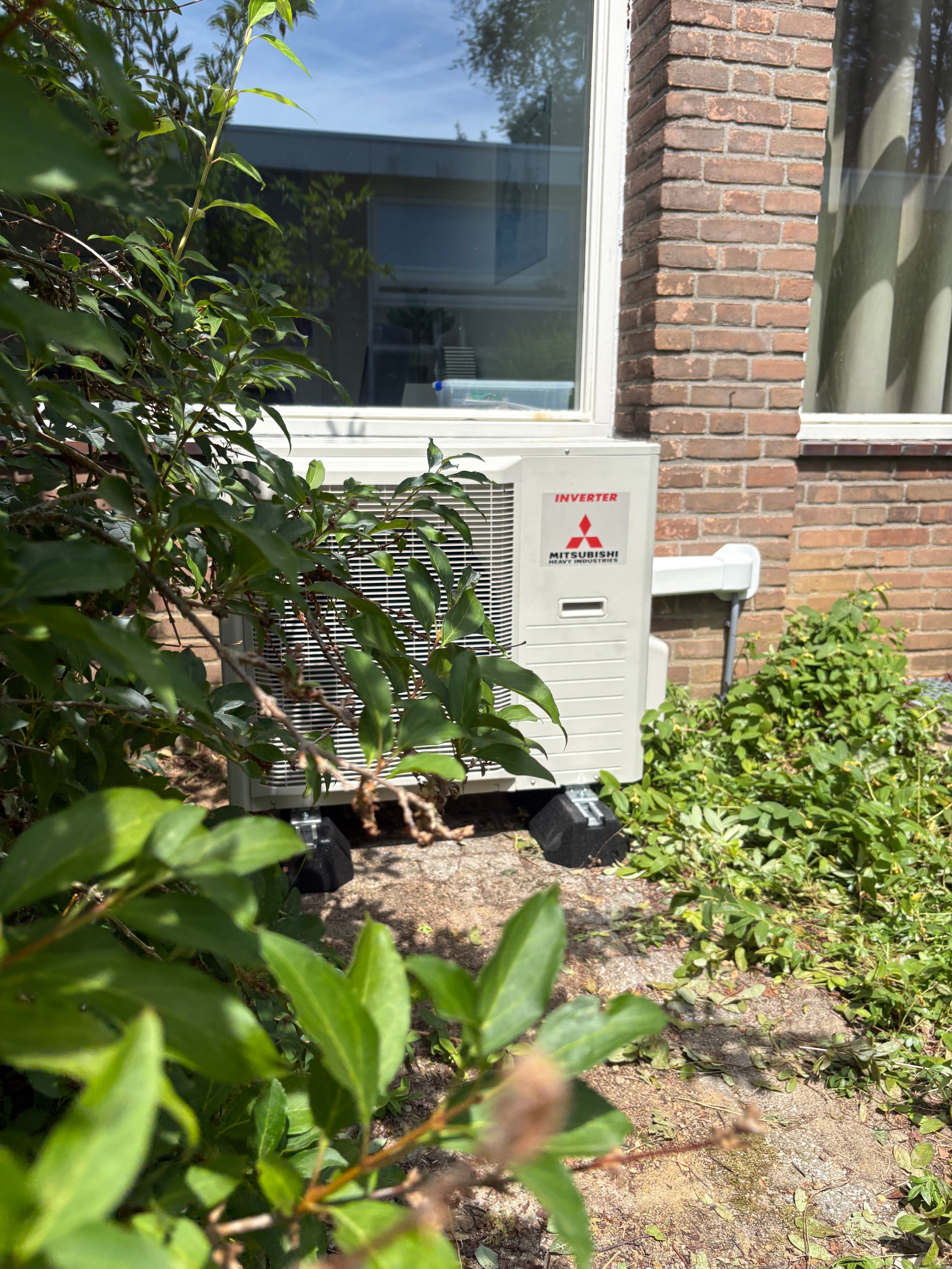 Mitsubishi heat pump unit installed against brick house wall, surrounded by green landscaping and foliage