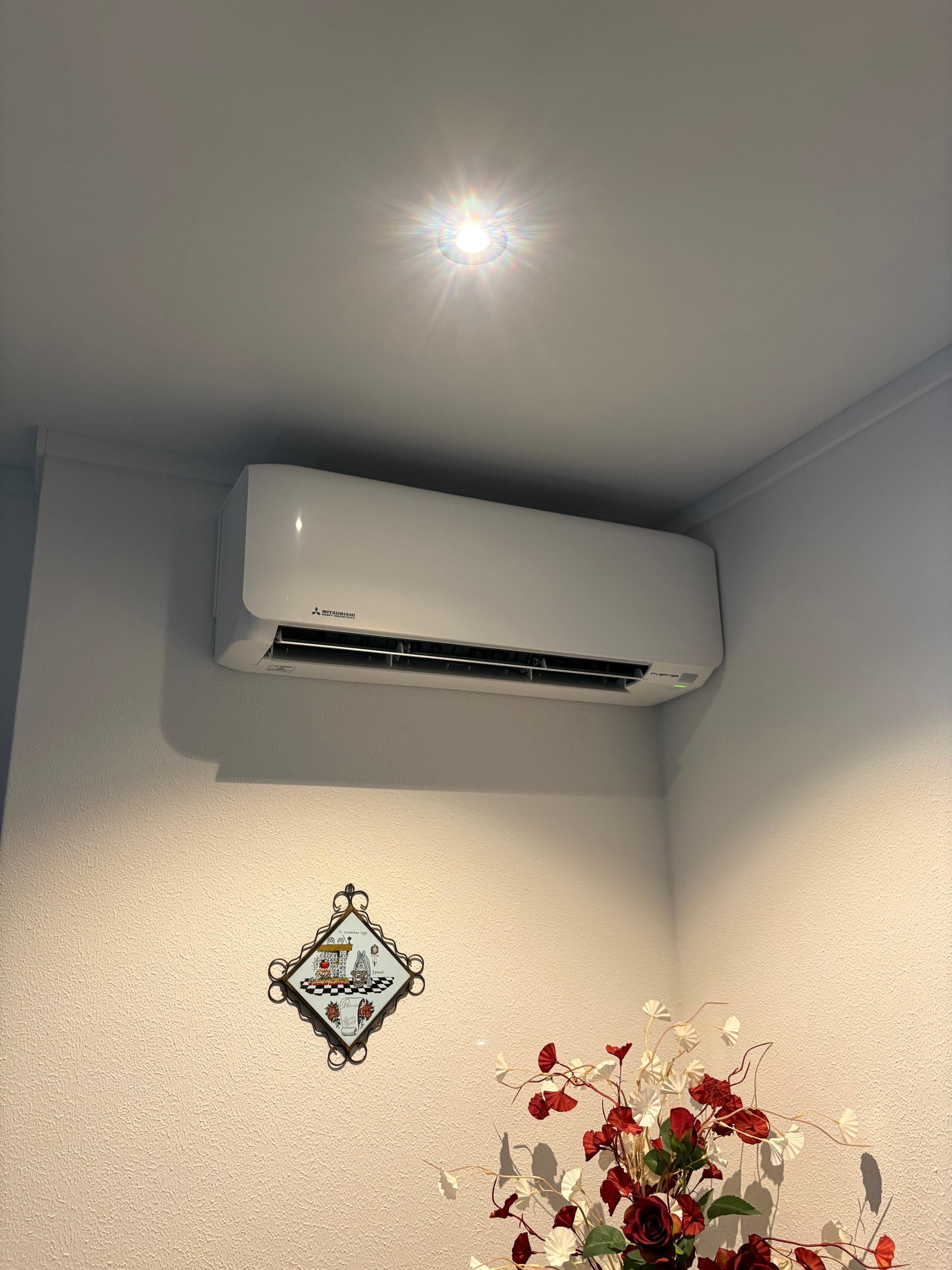 Indoor room corner with white split air conditioning unit on wall, decorative mirror, and red flowers in vase below