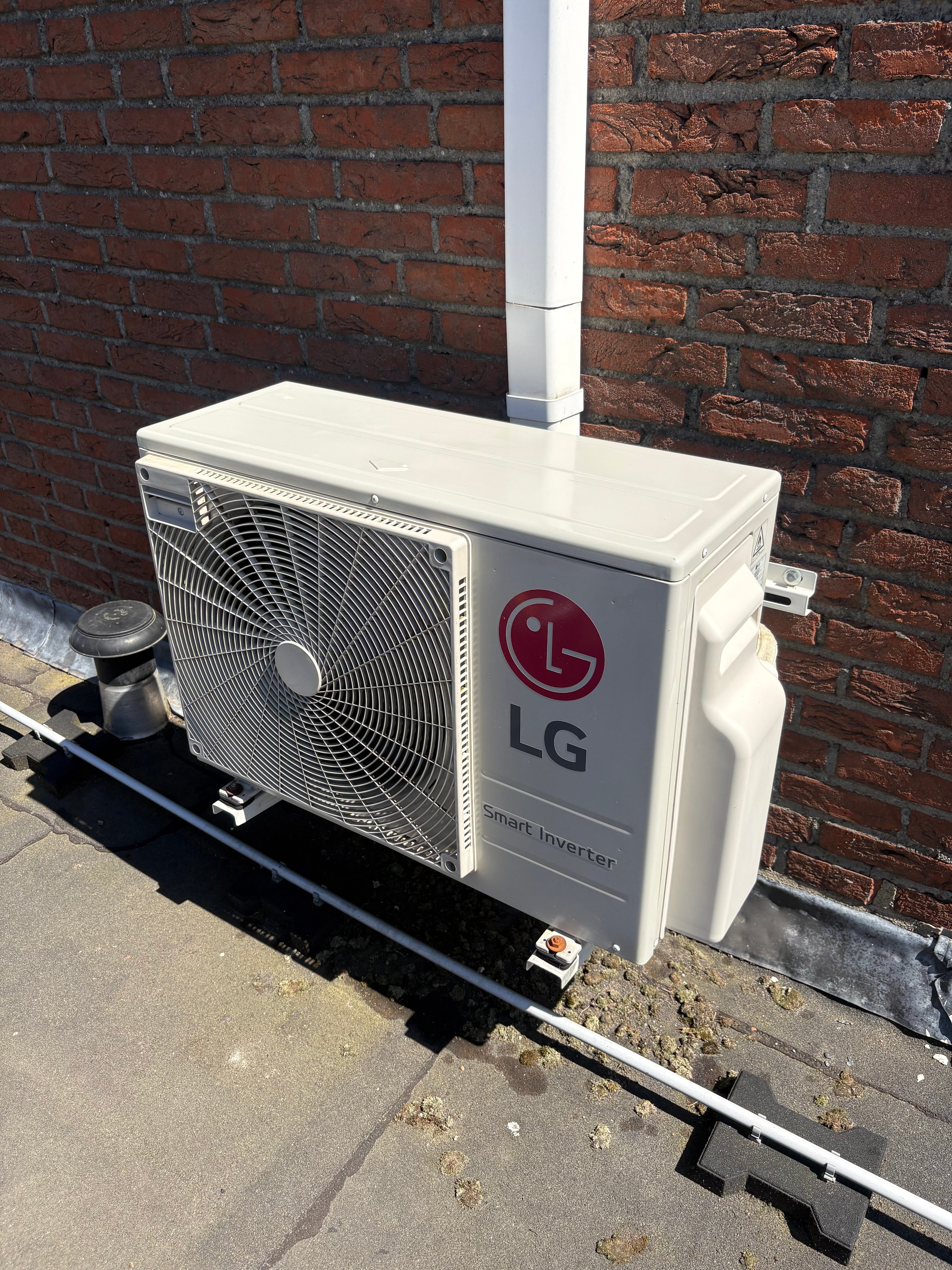 White LG air conditioning heat pump outdoor unit mounted against a red brick wall with white pipe