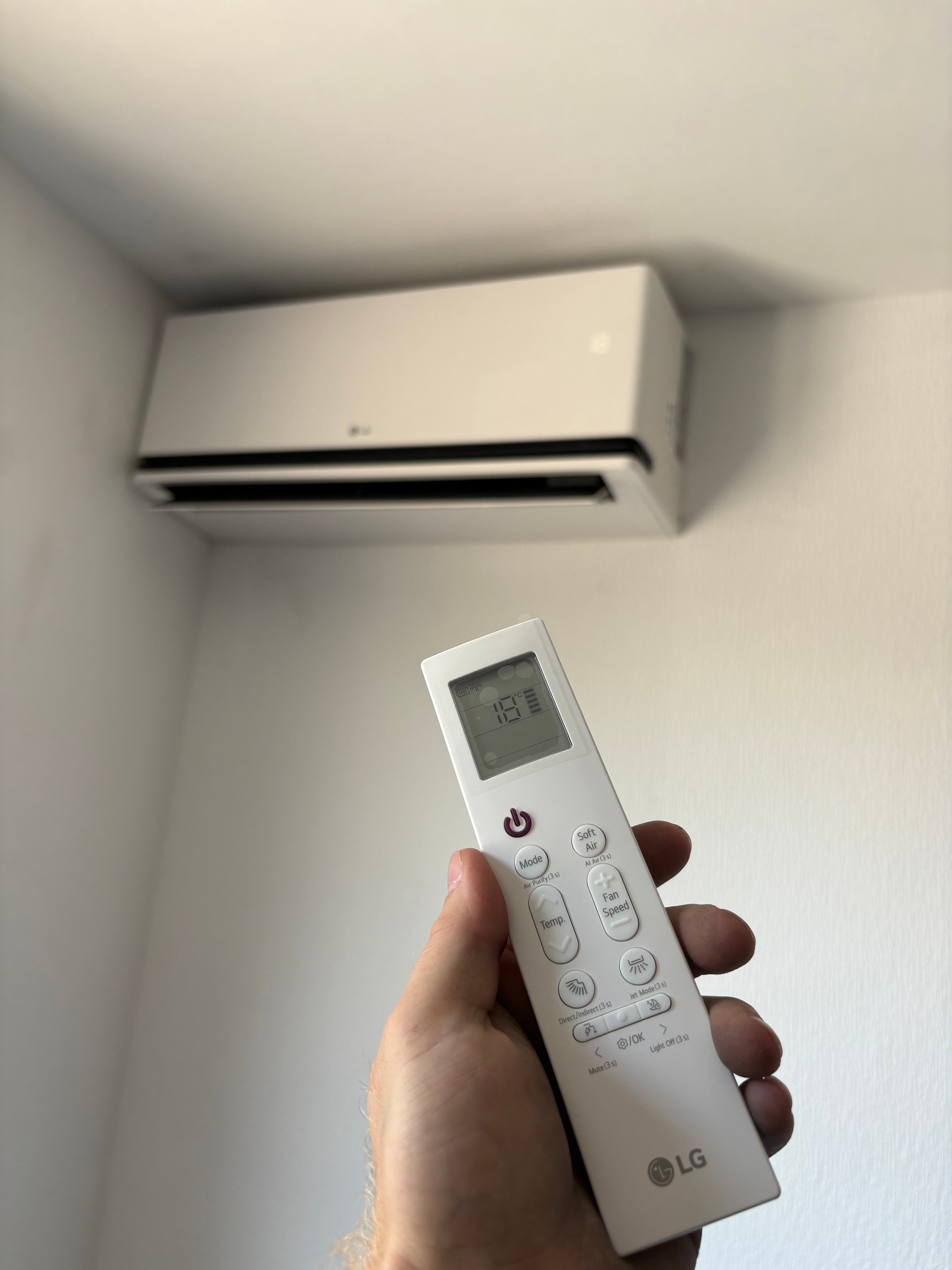 Hand holding white LG air conditioner remote with wall-mounted AC unit above