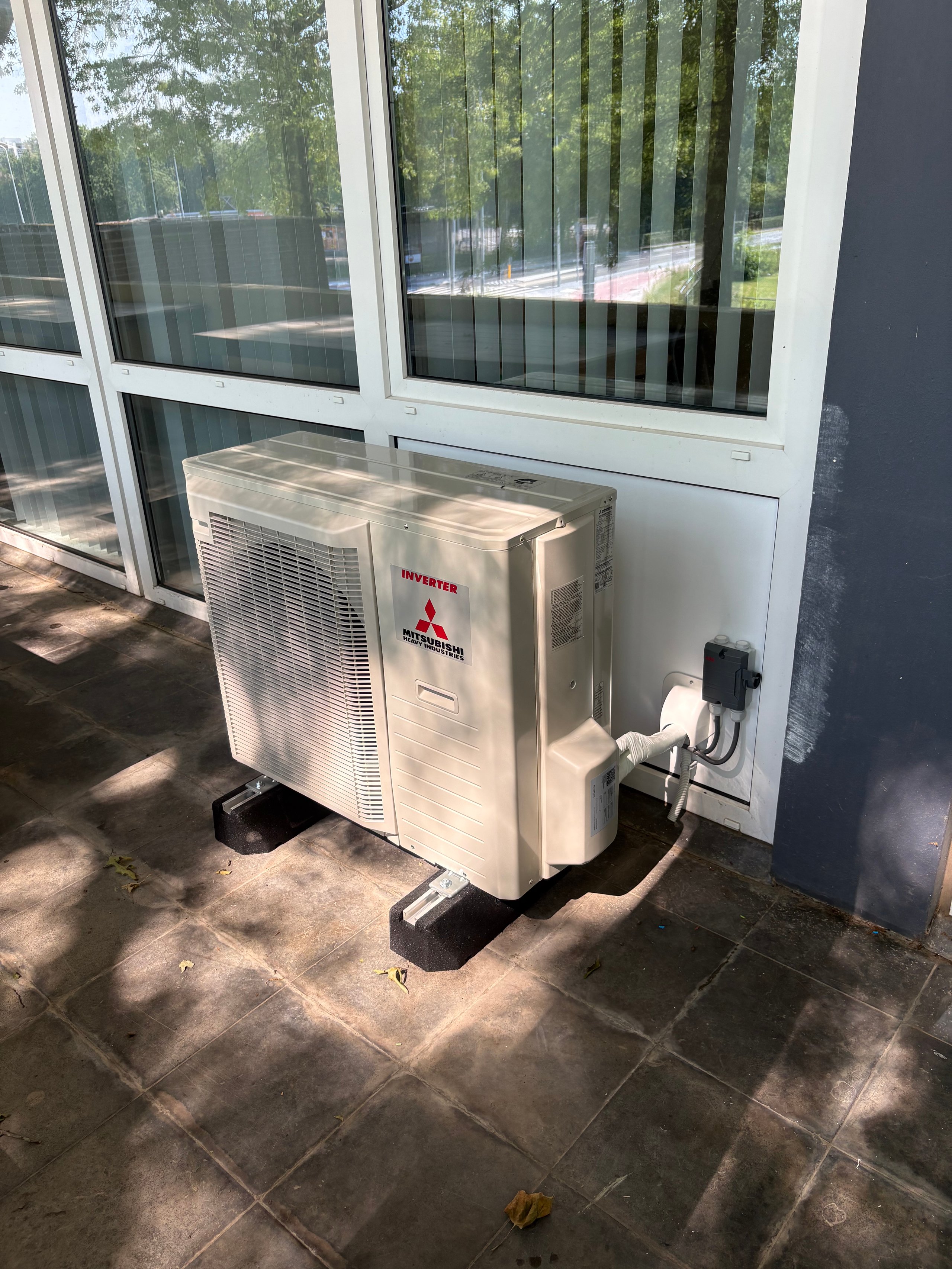 Mitsubishi air conditioning heat pump unit installed outside a residential home with glass doors and forest views