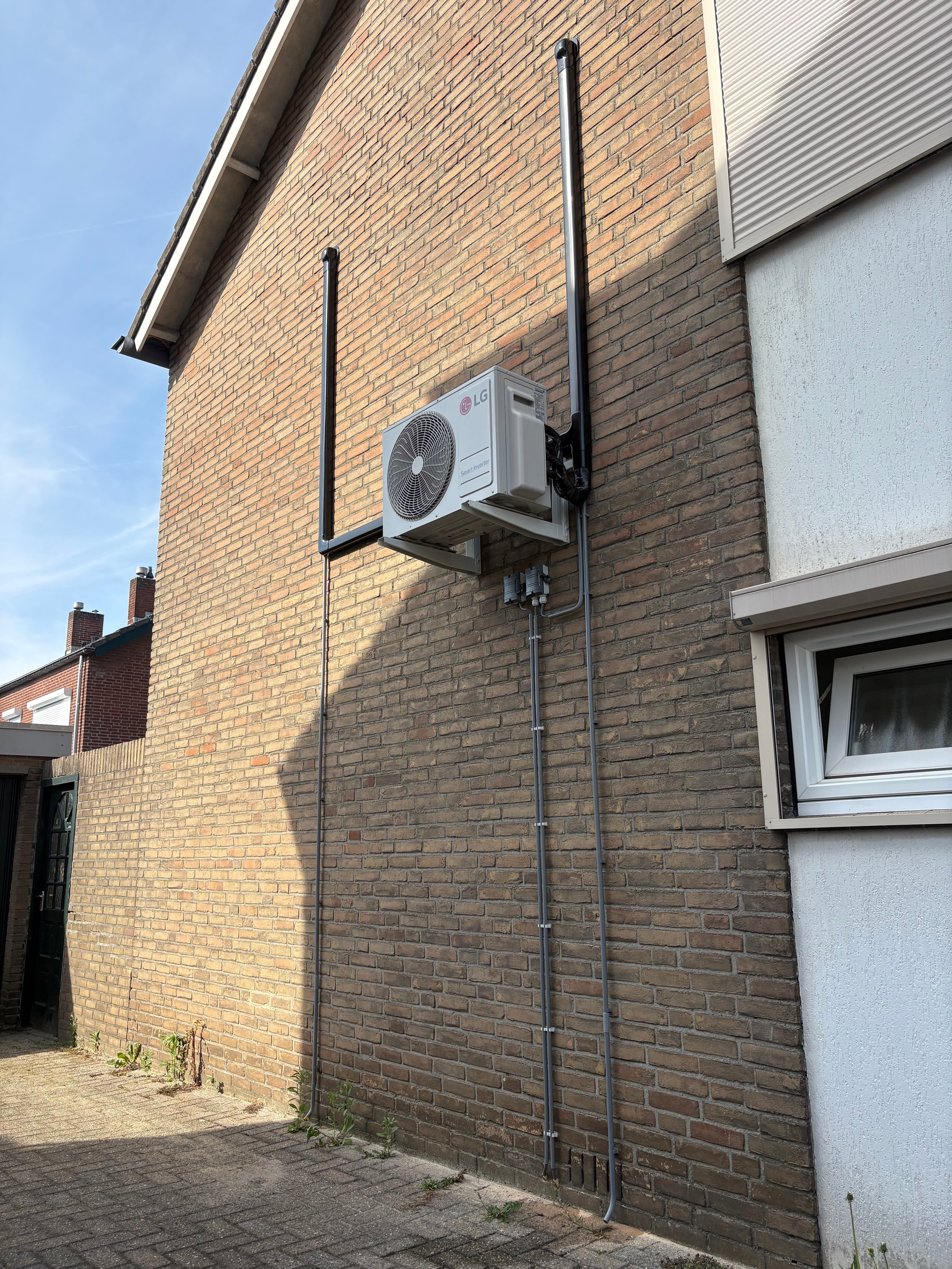 Outdoor heat pump unit mounted on brick building wall with black conduit pipes running vertically