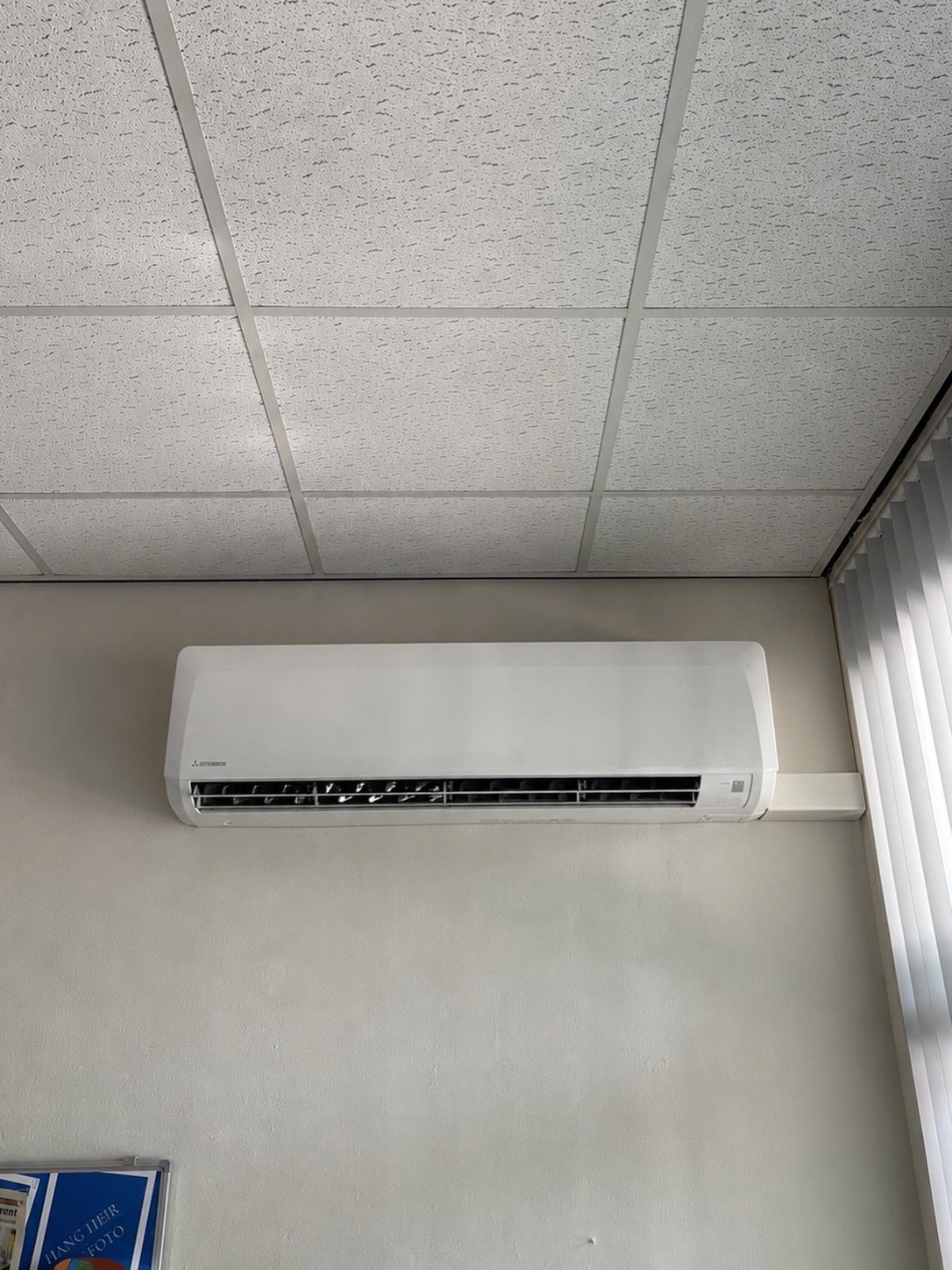 White split-type air conditioning unit mounted on a beige wall below a suspended ceiling with drop tiles.