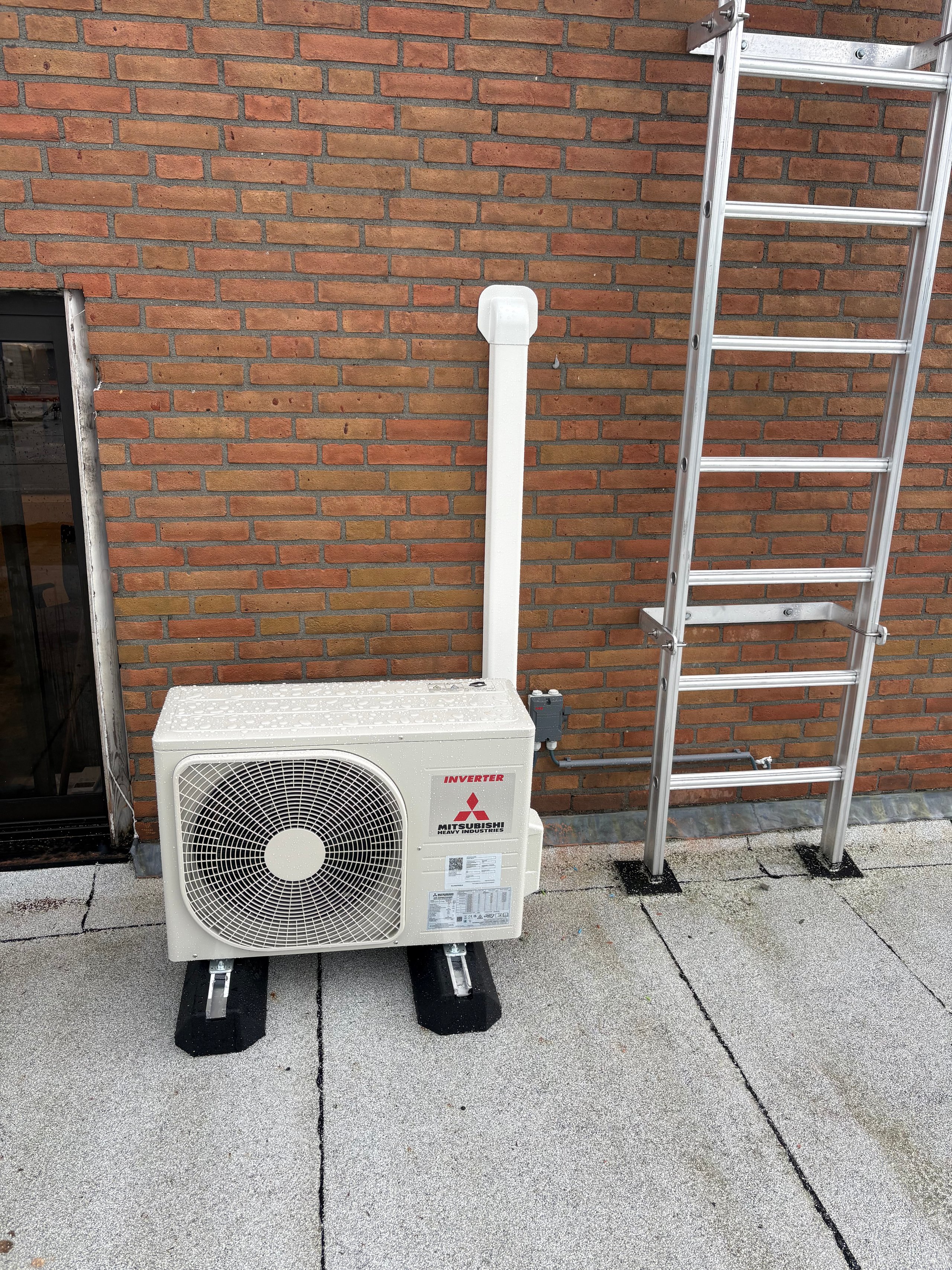 Mitsubishi air conditioning unit with white outdoor compressor and vertical pipe against red brick wall with aluminum ladder.
