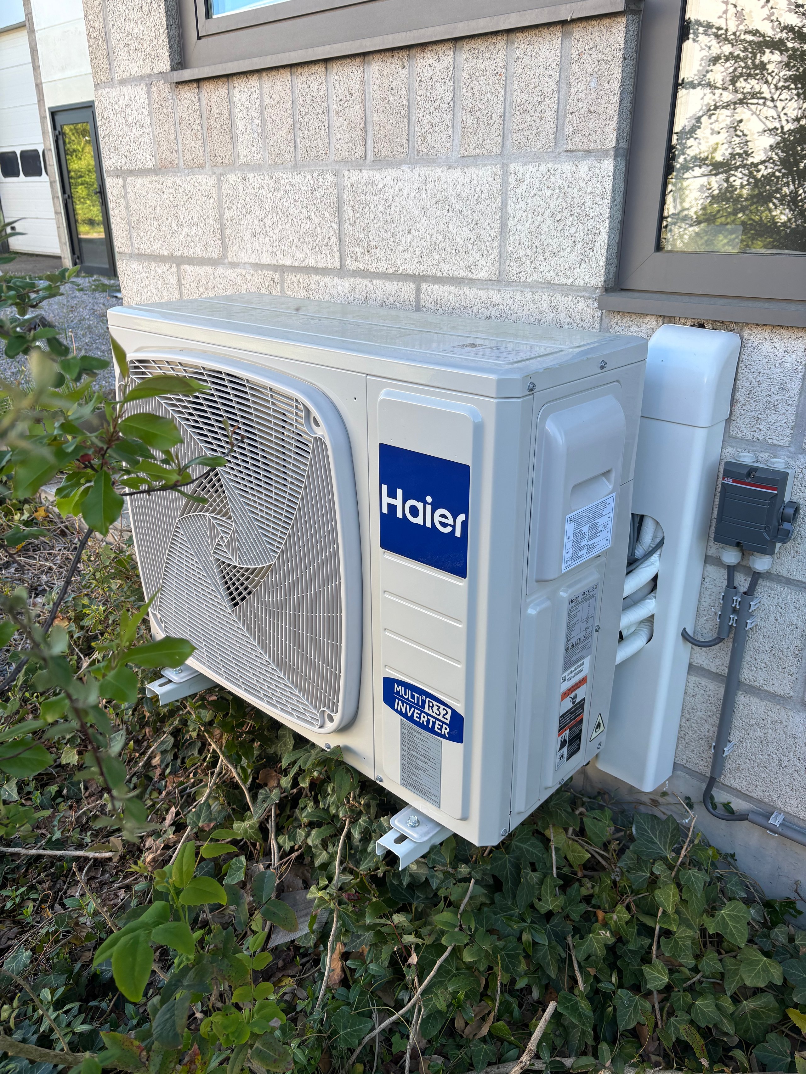 White Haier air conditioning outdoor compressor unit mounted on a building exterior with surrounding vegetation