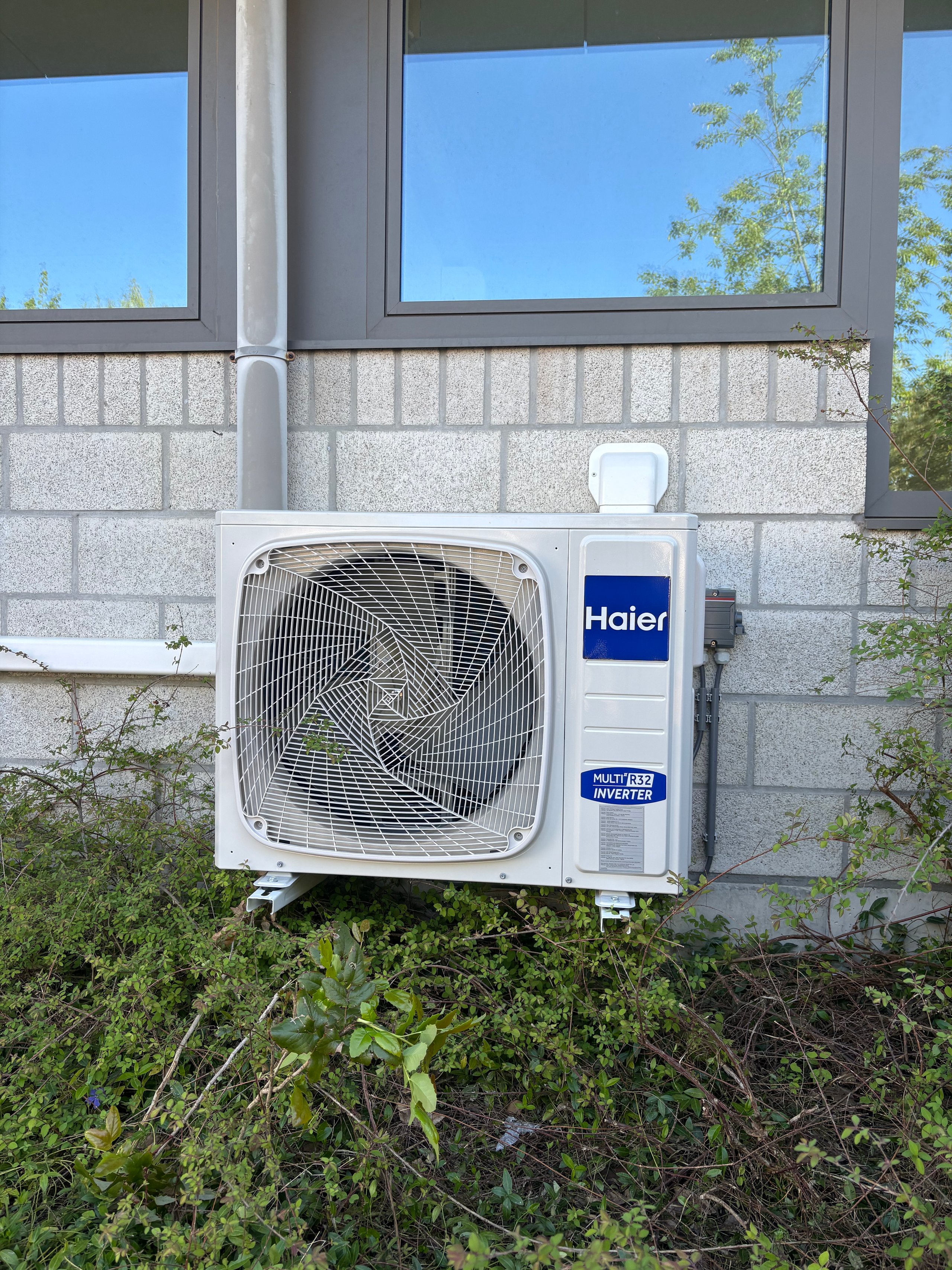 White Haier heat pump unit mounted on gray concrete block wall beneath windows with surrounding vegetation