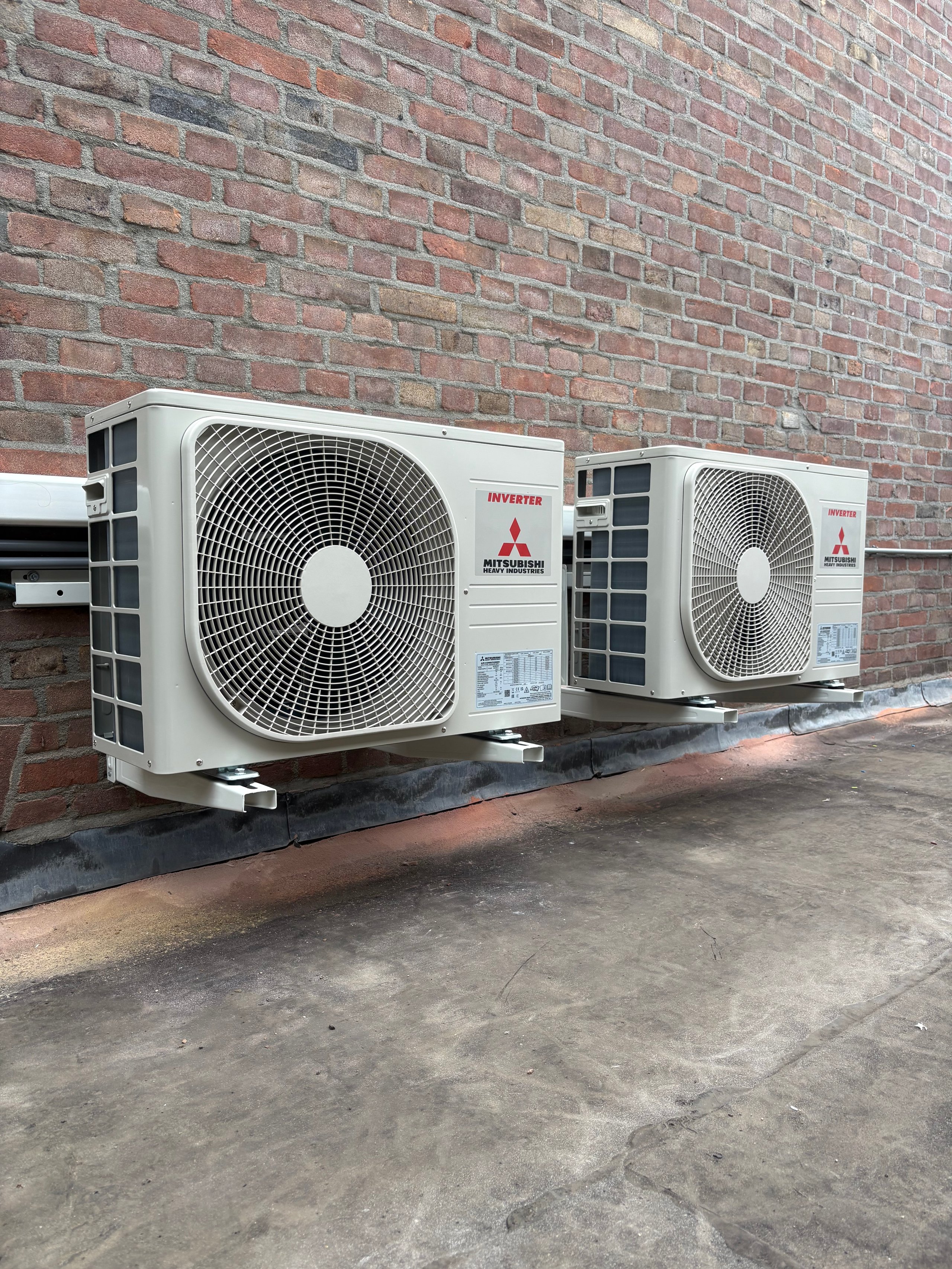 Two white Mitsubishi outdoor air conditioning units mounted on a brick wall with concrete foundation