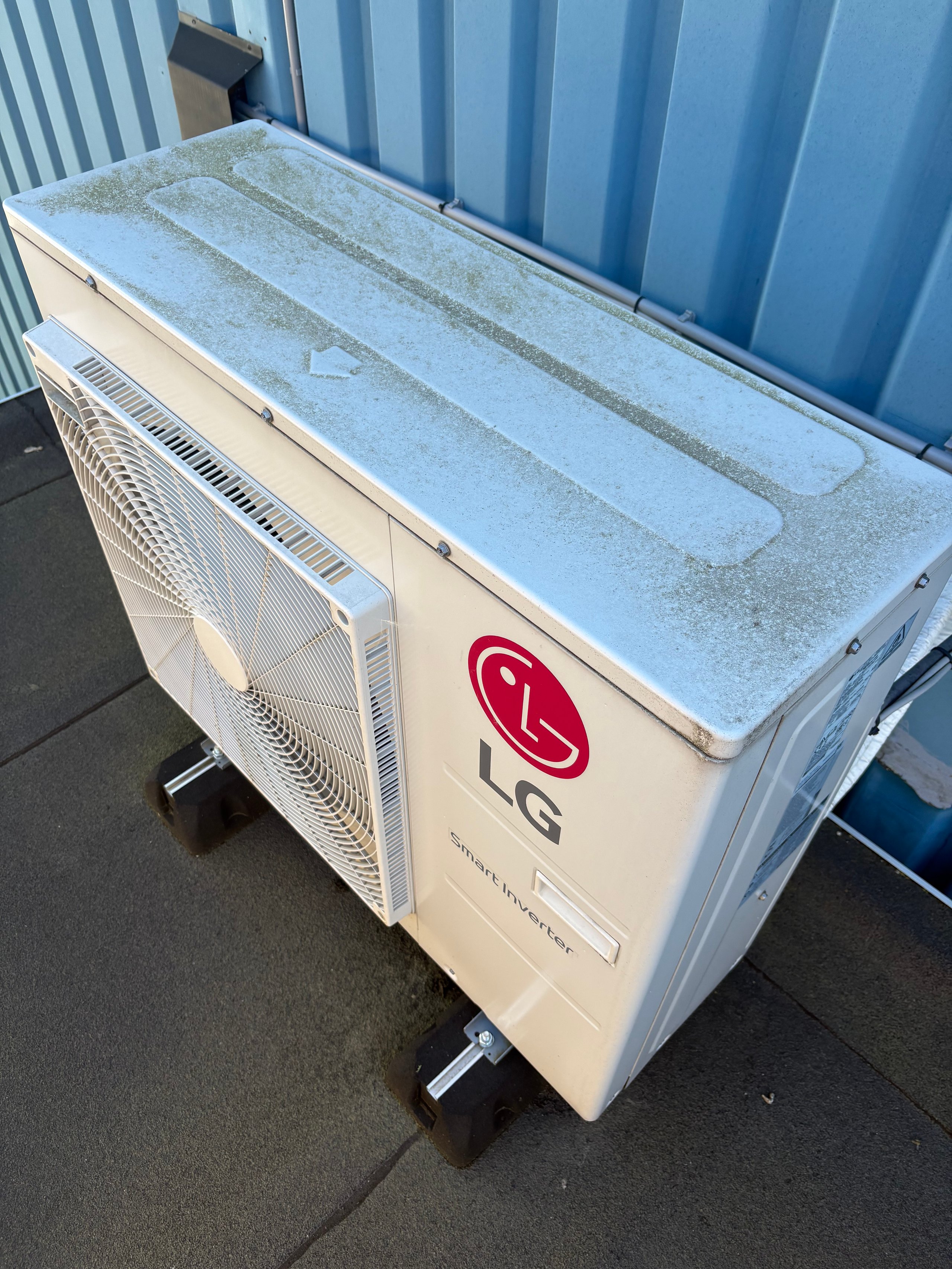 LG air conditioning outdoor unit with blue coolant lines and white housing on ground against blue corrugated metal wall